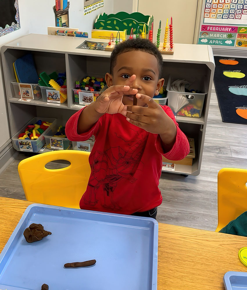 Toddler in a red shirt plays with brown playdough in a bright and organized classroom.