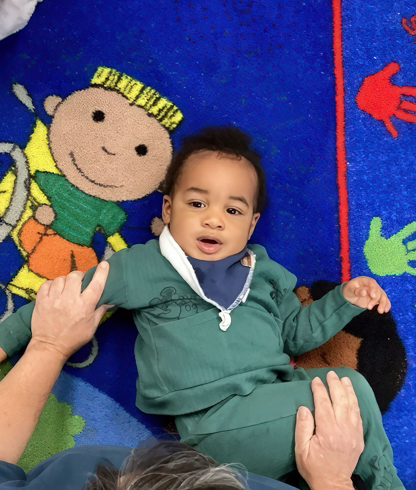 A baby wearing a green outfit lays on the floor, holding a person's hand, symbolizing warmth and affection