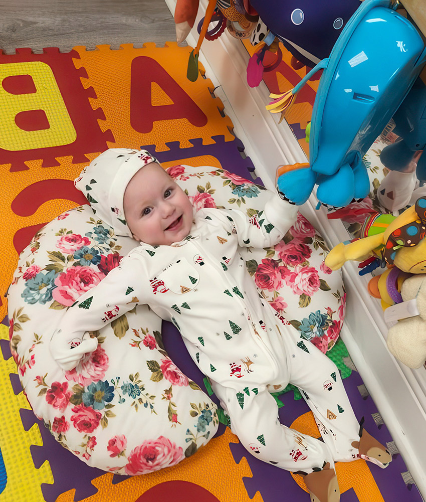 A baby dressed in a floral outfit lies peacefully on a colorful play mat, surrounded by soft toys and bright patterns.